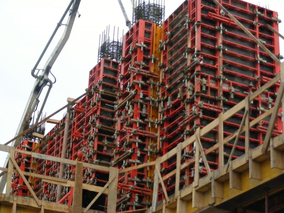 Cofraje metalice roșii montate pe pereții din beton armat ai unei clădiri în execuție, cu armătura ieșind deasupra și brațul pompei de beton în stânga