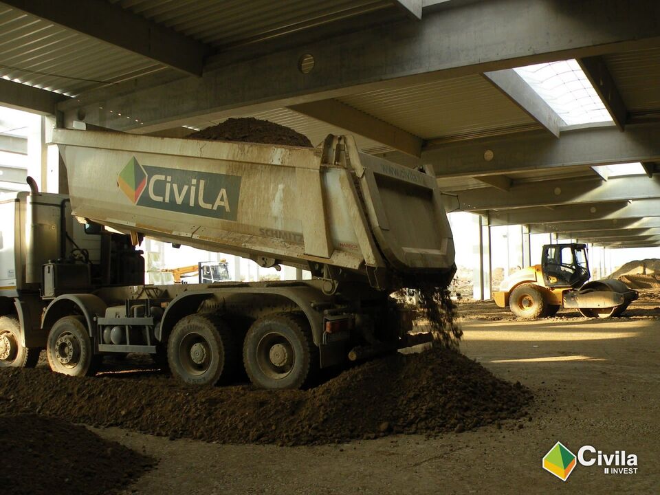 Basculantă Civila descărcând umplutură într-o hală în construcție, cu un compactor în spate
