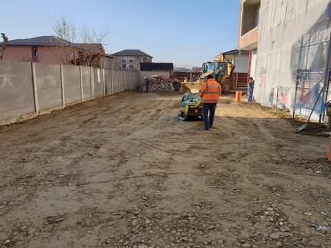 Compactarea curții blocului P+3 cu placa compactoare, cu buldoexcavatorul în spate și fațada acoperită cu plasă