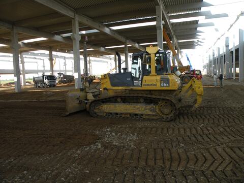 Buldozer la nivelarea umpluturii într-o hală cu structură prefabricată, cu basculante în fundal