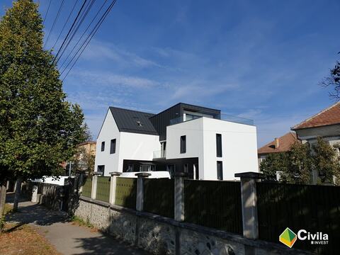 Casa particulară din Sibiu, finalizată: volume albe, acoperiș și placaj din tablă închisă la culoare, terasă cu parapet de sticlă, văzută din stradă peste gard