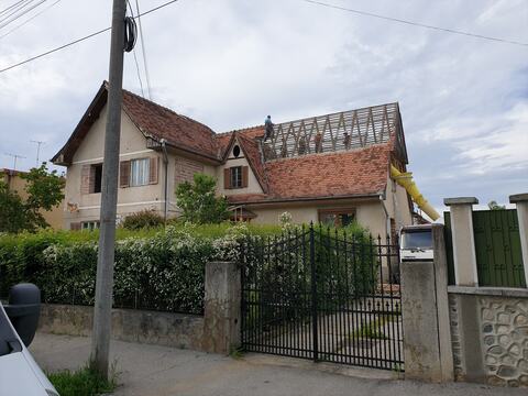 Acoperișul casei vechi în desfacere: țigla dată jos pe o parte, lații la vedere, muncitori pe acoperiș și tubul pentru moloz