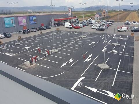 Parcarea centrului comercial din Târgu Secuiesc văzută de pe acoperiș, cu locurile și săgețile marcate; în fundal, un hipermarket vecin și munții