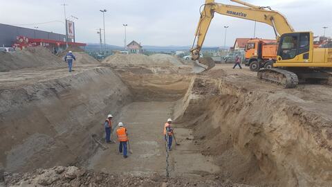 Săpătură adâncă, cu taluzuri, la Târgu Secuiesc: muncitori care curăță fundul săpăturii și excavatorul pe margine